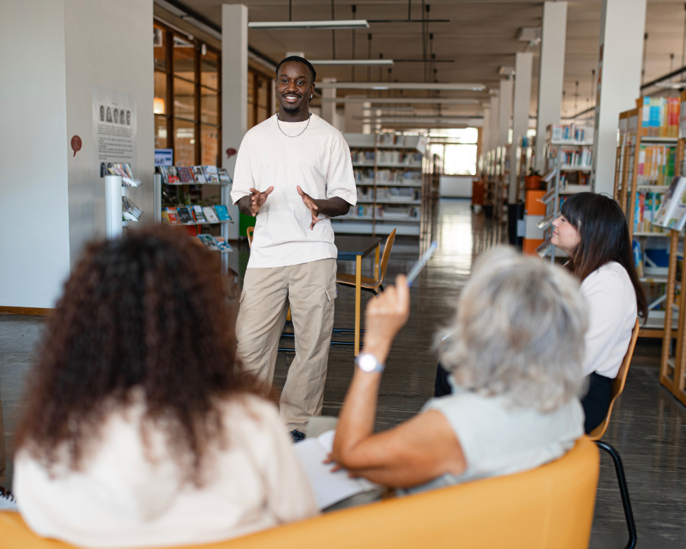 Young man leading a discussion in a library with seated adults listening.