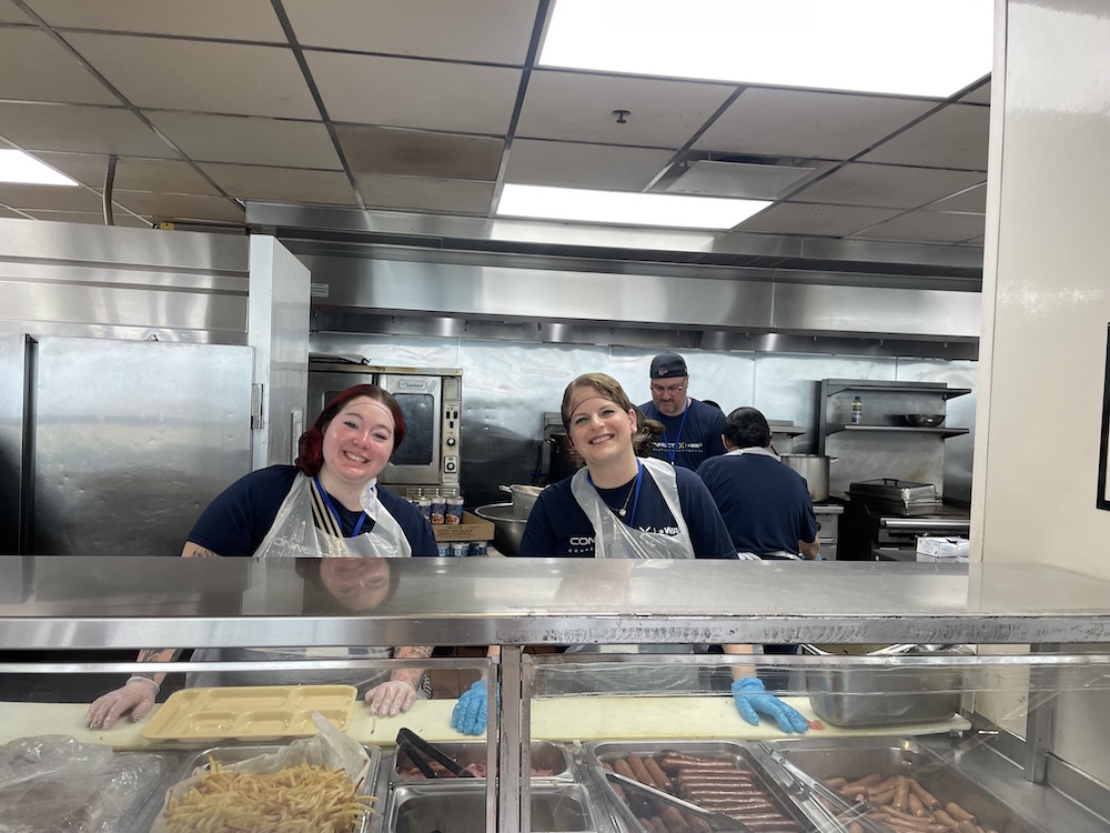 Volunteers smiling while serving food behind a kitchen counter.