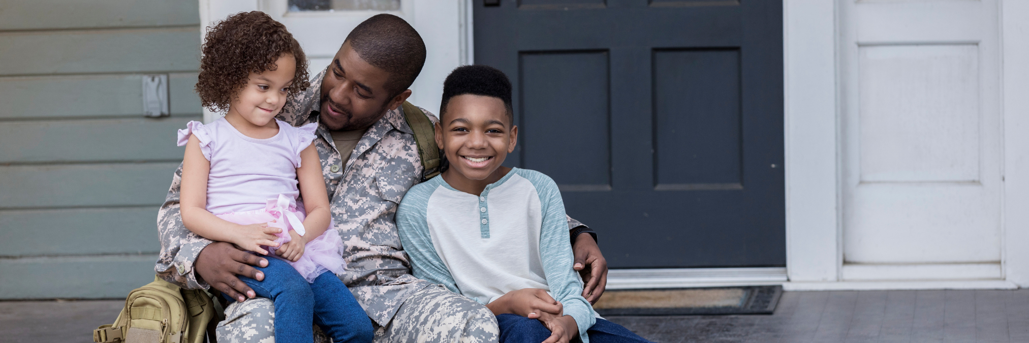 A U.S. military veteran sits on the front steps of a home, smiling with his two young children, symbolizing family, stability, and hope.