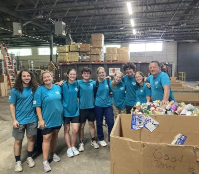 Group of volunteers in blue shirts posing inside a warehouse with boxes of canned goods