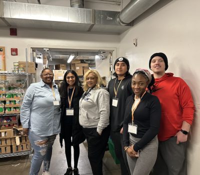 Six volunteers posing together in food pantry with shelves of food behind them