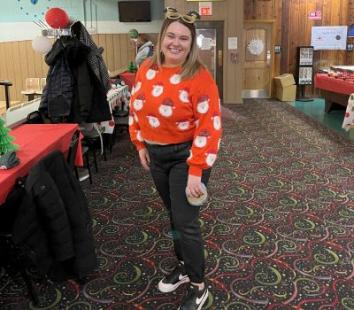 Woman wearing a red Santa sweater and holiday headband smiling in a decorated room