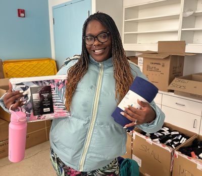 Smiling woman holding donation items including a water bottle, blanket, and hygiene kit