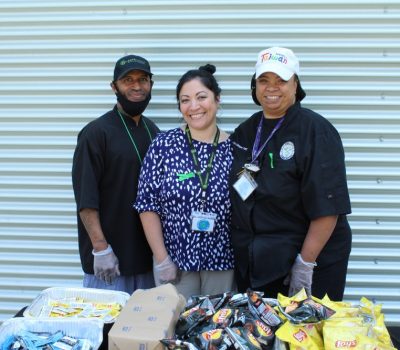 Three people smiling behind a table of snacks outdoors