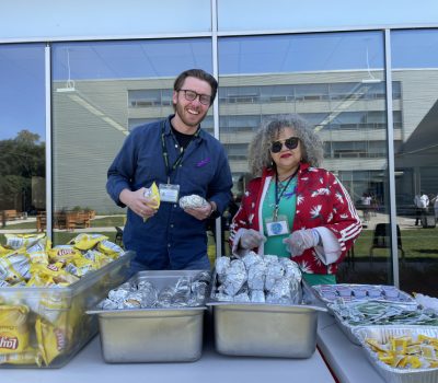Two team members serve pre-wrapped sandwiches, chips, and condiments outdoors at a food service table, smiling and ready to help.